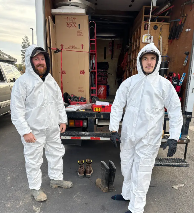 Two plumbers in their protective white garb, standing infront of their open work truck filled with tools